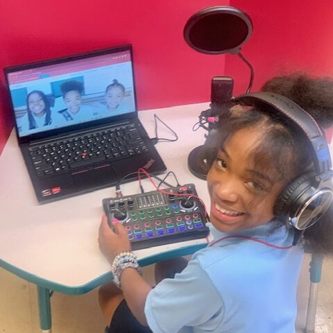 5th grade student wearing headphones, sitting in front of podcast equipment, smiling